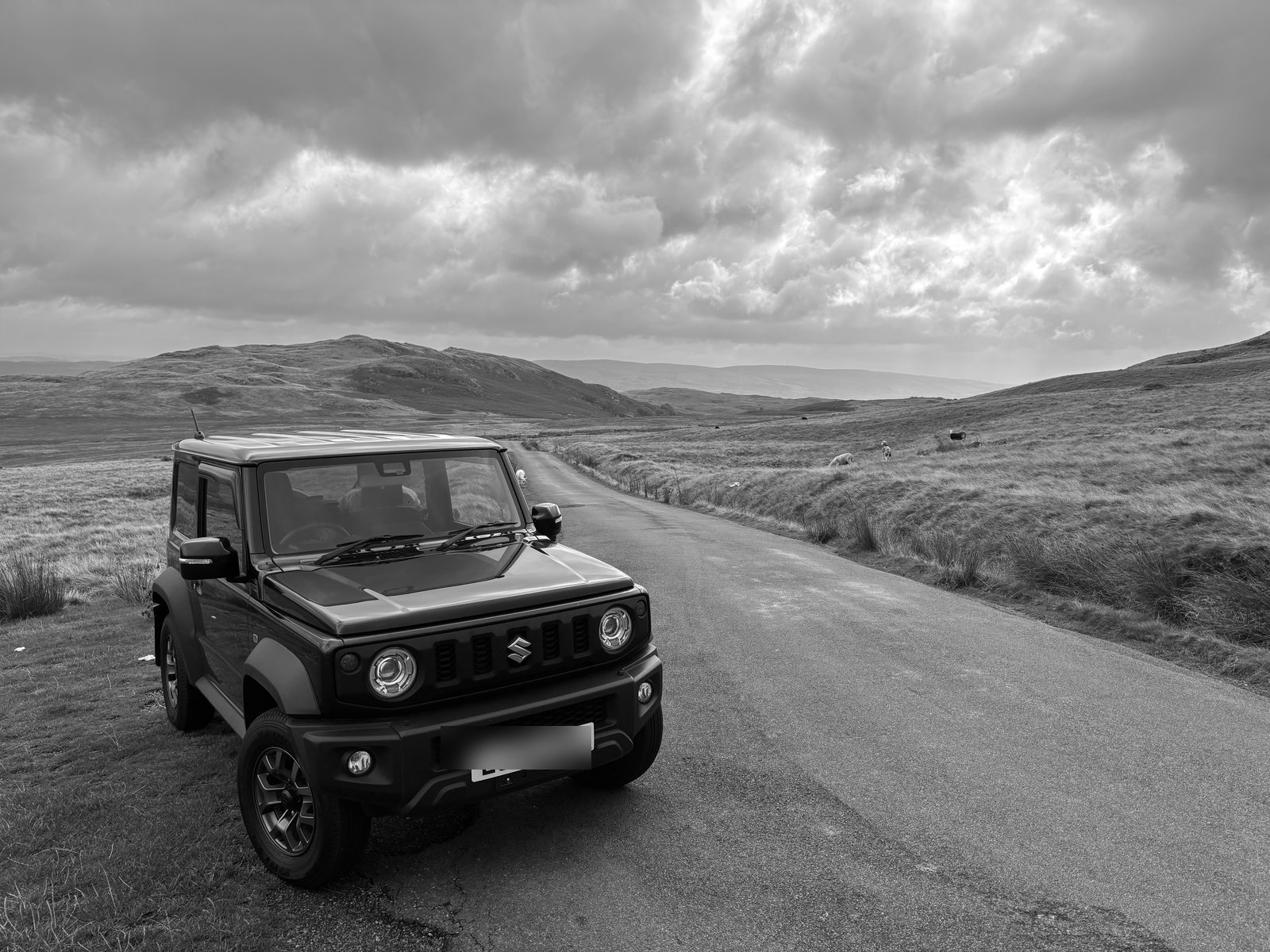 Black Suzuki Jimny parked on a hillside road with dramatic clouds overhead